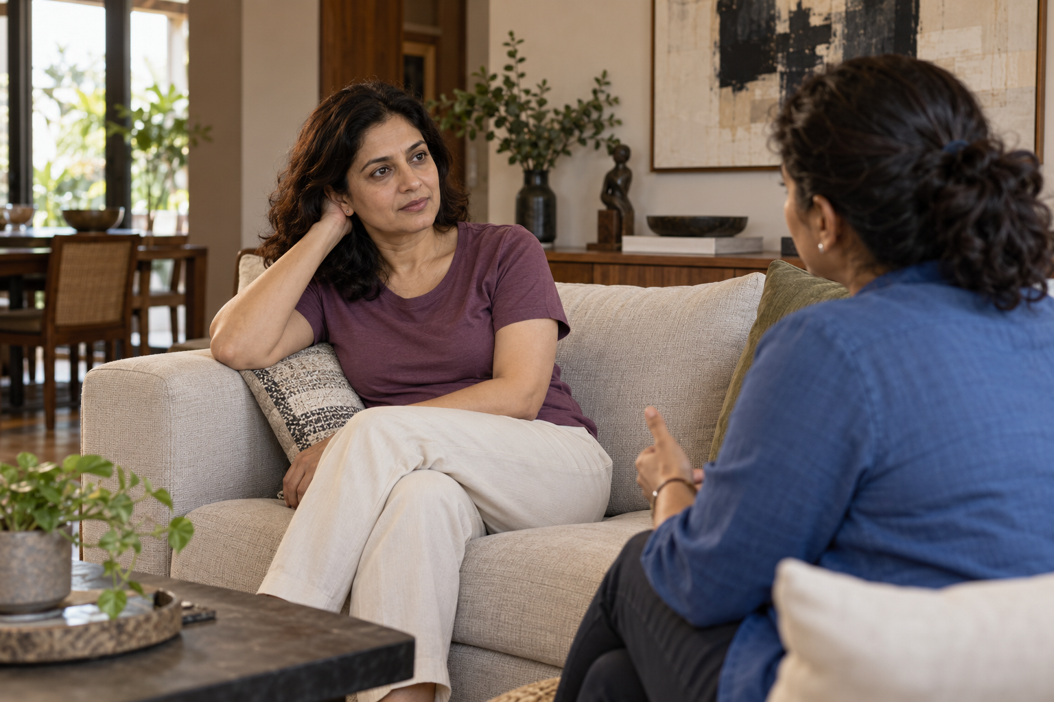 A woman in a plum T-shirt listening to a friend in a blue shirt in a sunlit apartment lounge.